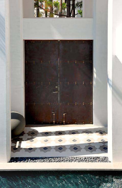 Industrial metal doors are closed behind white and gray Moroccan style patio tiles, while inset river rocks line an in ground swimming pool.