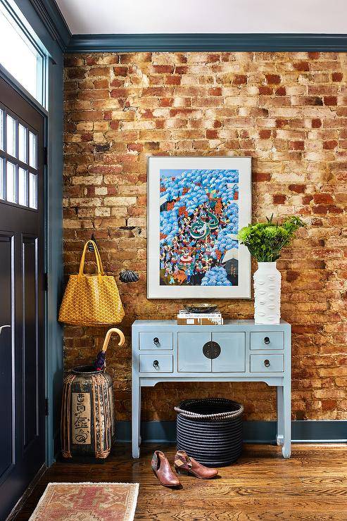 The eclectic foyer features a blue chinoiserie console table against an exposed brick wall with blue crown moldings and baseboards. The console is styled with wall art and a lips vase bringing a custom, eye-catching appeal.