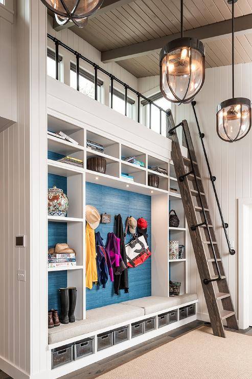 Cottage style mudroom features a brown oak ladder that leads to a loft with pipe plumbing rails, a mudroom locker with shelves and blue grasscloth wallpaper, a built in mudroom bench with metal storage show bins and a tan rug lit by vintage industrial light pendants.