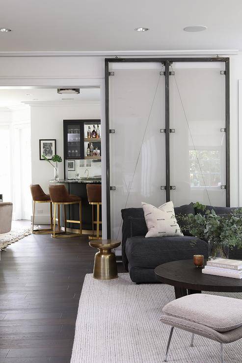 Chic living room boasts an industrial metal and glass sliding door on rails positioned behind a black comfy sofa paired with a gold accent table and positioned on a gray rug facing a round black coffee table.