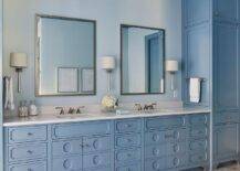 Glam blue bathroom features a gray marble floor tiles leading to a blue double washstand accented with ornate door fronts and a white quartz countertop. The washstand is fixed in a nook between blue built-in linen cabinets and beneath vanity mirrors hung from a light blue wall.