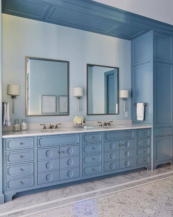 Glam blue bathroom features a gray marble floor tiles leading to a blue double washstand accented with ornate door fronts and a white quartz countertop. The washstand is fixed in a nook between blue built-in linen cabinets and beneath vanity mirrors hung from a light blue wall.