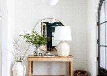 A black arched front door finished with glass panels opens to an entryway lit by a white glass and bronze lantern hung above a vintage red and black rug placed on a wood floor in front of a reclaimed wood console table. The table is topped with a large white lamp and located beneath a round mirror hung from a white painted brick wall.