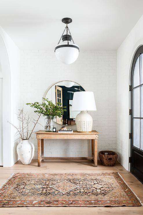 A black arched front door finished with glass panels opens to an entryway lit by a white glass and bronze lantern hung above a vintage red and black rug placed on a wood floor in front of a reclaimed wood console table. The table is topped with a large white lamp and located beneath a round mirror hung from a white painted brick wall.