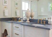 Glass and brass sconces are mounted over a full length frameless vanity mirror hung over antique brass hook and spout faucets mounted to a black honed marble countertop accenting a gray brushed oak dual washstand.