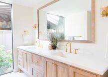A brown brushed oak dual washstand adorned with brass hardware is topped with a white quartz countertop holding brushed gold faucets beneath a brown brushed oak vanity mirror flanked by brass and glass sconces.