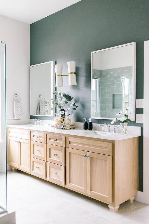 White framed vanity mirrors hang from a hunter green painted wall on either side of white and gold sconces fixed above a golden oak dual bath vanity.