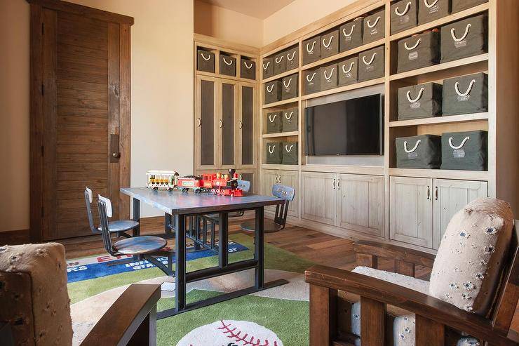 Cottage playroom room features a full wall lined with a distressed shelving unit and cabinets filled with gray canvas bins with white rope handles fitted with a flatscreen TV niche. Kids' homework room boasts an industrial desk with fitted with metal chairs atop a baseball themed rug.