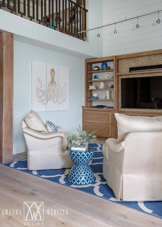 Brown and blue cottage living room clad in blond wood floors covered in a blue chain link rug features a 3 piece octopus art piece mounted on a light blue wall and a two inward facing tan silk skirted chairs separated by a blue hourglass stool and facing shiplap accent well boasting a styled limed oak built in television shelving unit fitted with a flat panel television illuminated by a modern track light.