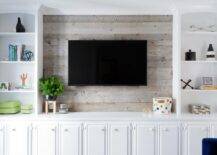 Styled white built in cabinets flank a barn board wall fitted with a flat panel television mounted above white TV cabinets.