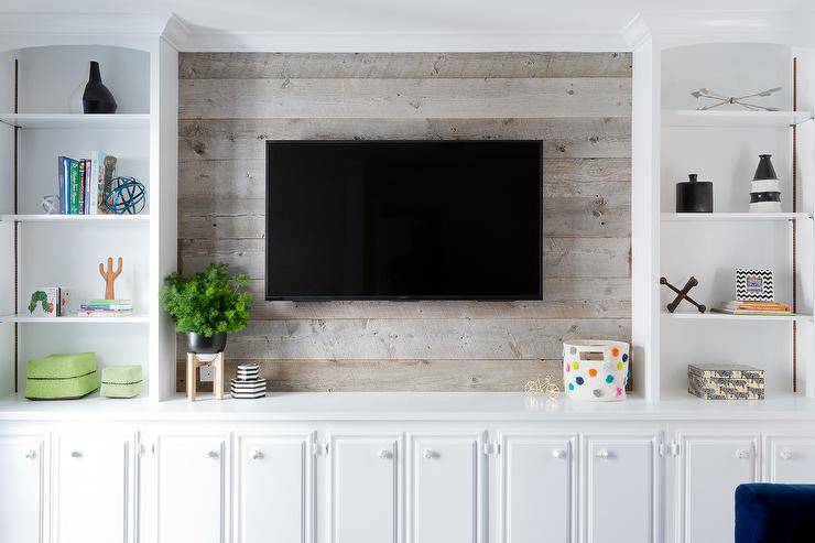 Styled white built in cabinets flank a barn board wall fitted with a flat panel television mounted above white TV cabinets.