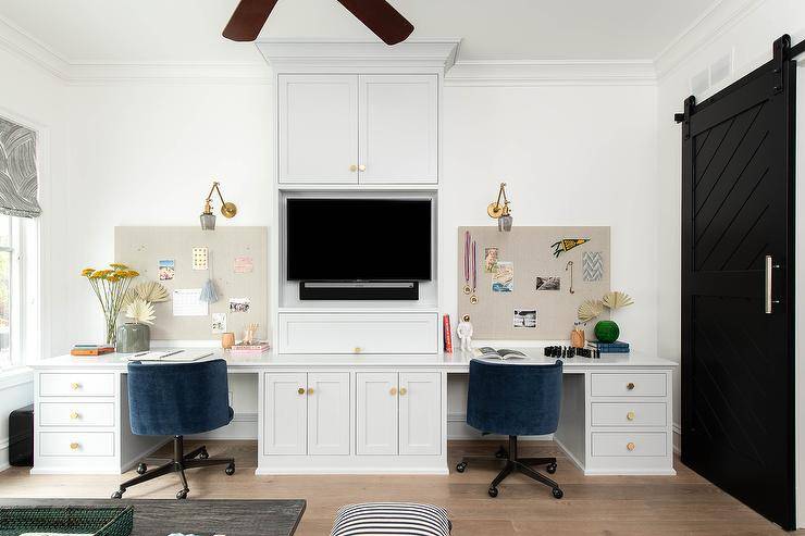 A black barn door on rails opens to a family room boasting light gray built-in desks painted in Benjamin Moore Graytint, accented with brass hexagon knobs, and seating blue desk chairs facing pin boards lit by brass swing arm sconces. The desks flank light gray built-in cabinets holding a flat panel television.