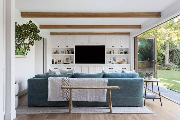 A sawhorse bench sits at the back of a blue sectional placed beneath a ceiling fitted with stained wood beams facing a white custom built-in TV unit accented with oil rubbed bronze hardware and styled white shelves. Natural lights stream in front folding glass patio doors.