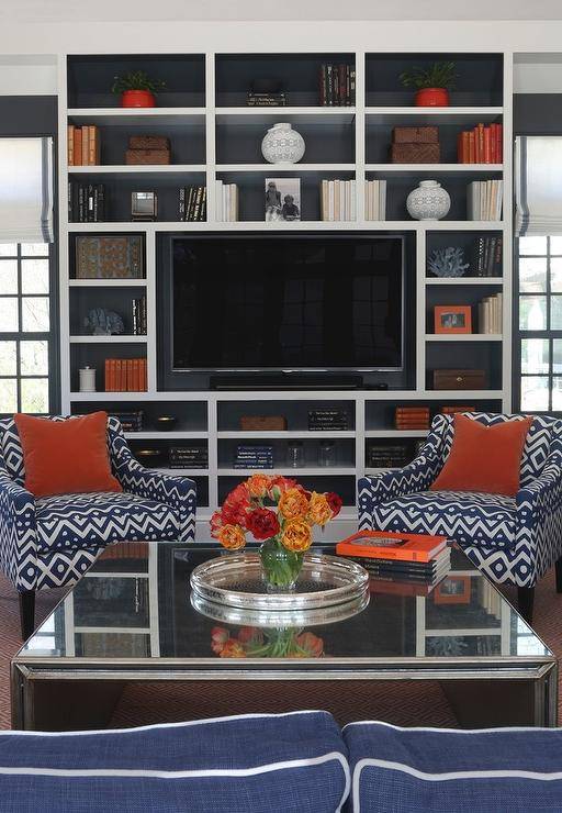 Blue and orange living room features a mirrored waterfall cocktail table and a pair of blue geometric accent chairs lined with orange velvet pillows placed before floor to ceiling built in media unit, with backs of shelves painted blue, filled with a flatscreen TV and orange accents.
