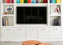 A white built-in shelving media unit not only looks great but is a practical space saving design. Closed-door storage hides the unattractive components, wires, or anything you wouldn't want on display. Open bookshelves and cubbies boast colorful decor and books creating a bubbly contrast to the white shelving finish. A flat-panel tv is the center of it all over a set of bottom drawers with lucite and black knobs. A gold metal coffee table energizes the view over a white round shag rug layered over gray washed wood floors.