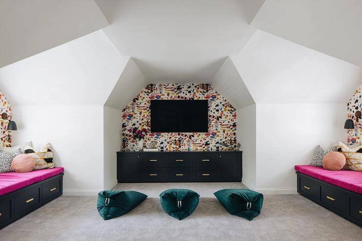 Stylish attic playroom features a wall mount TV fixed in a nook to an accent wall covered in paint splatter wallpaper over a black built-in TV unit. Three peacock blue bean bags sit on gray carpet between facing black built-in window seats finished with drawers and pink cushions.
