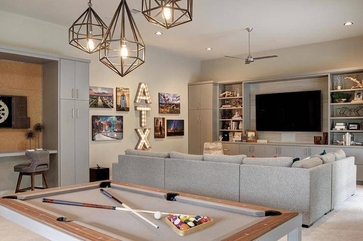 Staggered polyhedron pendants are hung over a pool table positioned behind a gray sectional facing a gray television unit fitted with gray cabinets and styled gray shelves flanking a flat panel television. Marquee lights are mounted to a gray wall between canvas photographs and near a gray built in desk boasting gray cabinets surrounding a cork board backsplash.