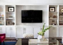Transitional living room features a wall to wall white built in shelving unit lined with a flat screen TV and grey trellis wallpaper on the back of shelves.