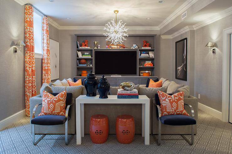 Orange and gray basement family room features walls clad in gray grasscloth wallpaper lined with a window dressed in orange ikat curtains in Quadrille Island Ikat Fabric Orange. Chic basement family room boasts a gray pit sectional adorned with navy and orange pillows illuminated by a Arteriors Diallo Small Chandelier facing a gray media cabinet filled with a flatscreen TV and orange foo dogs. A pair of orange stools is tucked below a West Elm parsons Console Table topped with navy ginger jars flanked by navy velvet chairs adorned with orange ikat pillows.