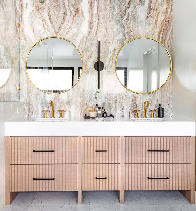 Modern bathroom features round brass mirrors mounted on a pink and gray marble slab accent wall over a beige reeded double washstand with brush gold faucets.