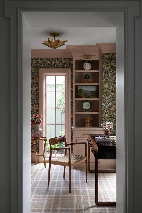 Home office features strawberry thief wallpaper, mauve built in shelves and a mid century modern desk chair at a vintage desk atop pink and gray plaid carpeting.