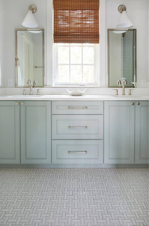 A bamboo roman shade hangs from a window flanked by nickel framed vanity mirrors lit by nickel sconces with white conical shades. The sconces light polished nickel gooseneck faucets fitted to a gray green dual washstand placed on marble weave floor tiles.