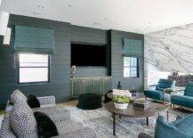 Turquoise blue roman shades hang from windows framed by hunter green shiplap and flank an inset TV fixed above turquoise blue built-in cabinets lined with gold trim. Side-by-side black and white chevron accent chairs and turquoise blue leather and brass chairs sit on either side of industrial nesting coffee tables placed on a gray rug.