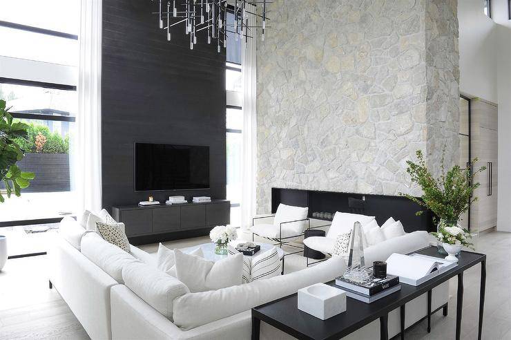 This luxurious black and white modern living room a black sofa table placed behind a white sectional topped with white pillows and paired with a round marble top coffee table. Two black sleek wire chairs flank a black accent table that complements a black floating tv unit positioned under a wall mounted tv fixed to a black accent wall. A gray stone 2-story fireplace is located adjacent to floor-to-ceiling windows.