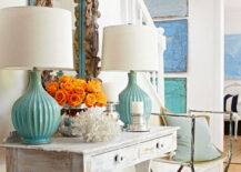 Pretty gray and turquoise entryway with beachy, coastal theme. A distressed whitewashed console table with drawers is paired with a driftwood framed mirror. A pair of glazed turquoise table lamps flank the mirror. Bright orange roses, a silver candle holder and piece of coral adorn the table. An antique iron chair is paired with a blue pillow. On the stairwell wall three blue canvases hang vertically.