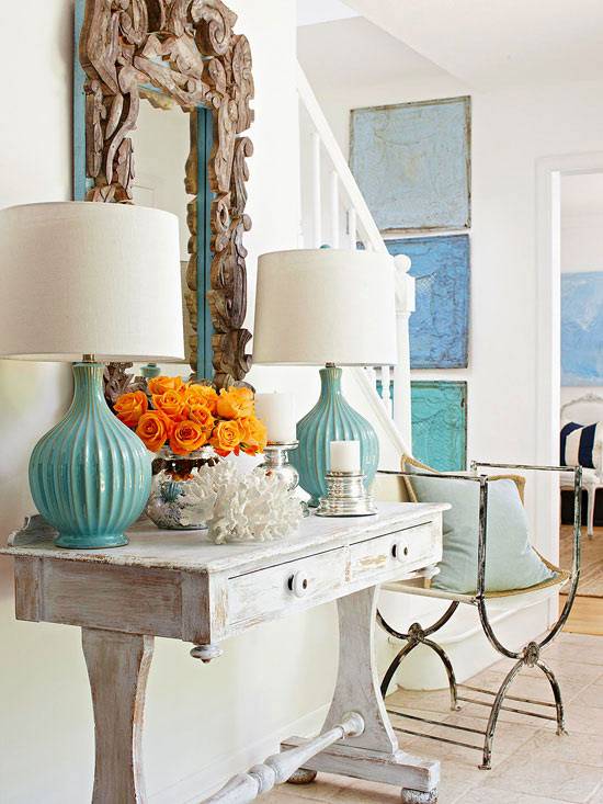 Pretty gray and turquoise entryway with beachy, coastal theme. A distressed whitewashed console table with drawers is paired with a driftwood framed mirror. A pair of glazed turquoise table lamps flank the mirror. Bright orange roses, a silver candle holder and piece of coral adorn the table. An antique iron chair is paired with a blue pillow. On the stairwell wall three blue canvases hang vertically.