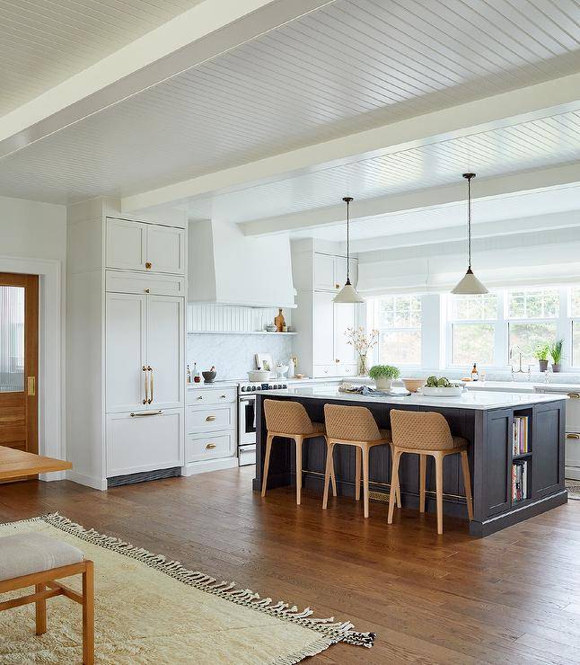 White u-shaped kitchen is contrasted with a black island fitted with cookbook shelves and seating brown woven stools at a white and gray marble countertop lit by two black and white vintage lanterns hung from a beadboard ceiling.