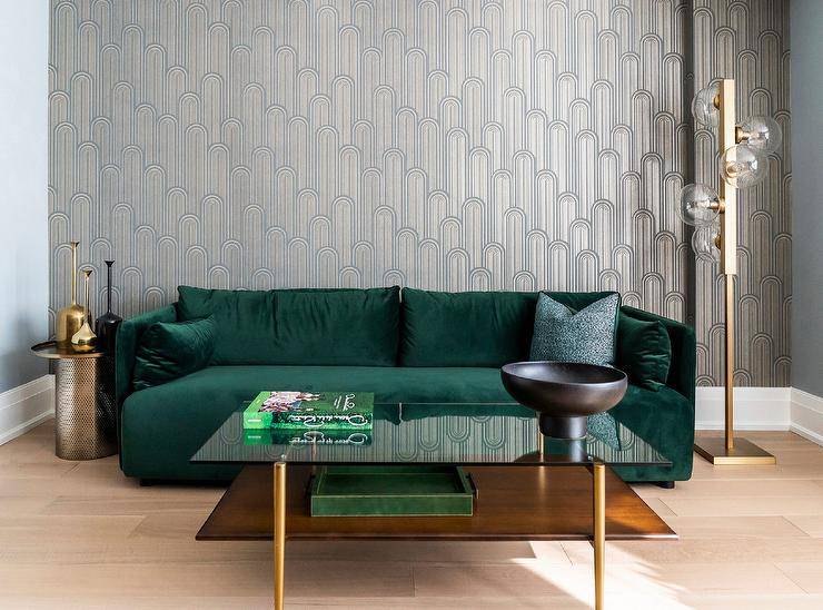 Chic Speakeasy Wallpaper covers an accent wall complementing blue walls and fixed behind an emerald green velvet sofa matched with a round brass end table and a black glass and brass coffee table.