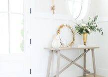 Gorgeous entryway features a styled gray wash wooden trestle table placed on a gray wash wide plank floor in front of a board and batten wall and beneath an oval mirror lit by a brass sconce.