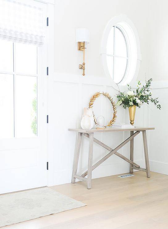 Gorgeous entryway features a styled gray wash wooden trestle table placed on a gray wash wide plank floor in front of a board and batten wall and beneath an oval mirror lit by a brass sconce.