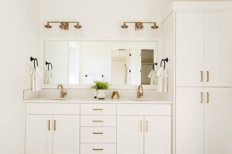 Off white washstand with brass hardware and white quartz featuring brushed gold faucets under a white wooden framed full-length vanity mirror illuminated by 3-light brass sconces.