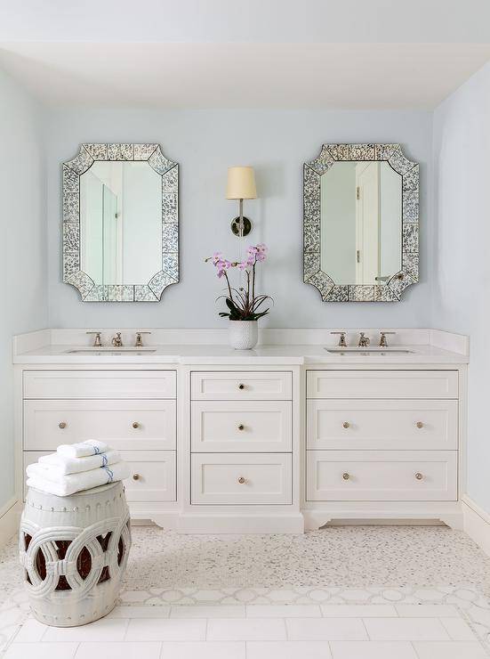 Two antiqued French mirrors hang on either side of a Camille Long Sconce over a white dual bath vanity adorned with polished nickel knobs and polished nickel faucets. A white rope stool sits on ivory mosaic floor tiles in front of the vanity.