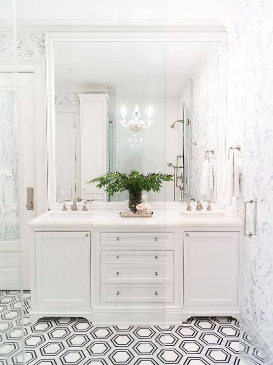 A white beveled mirror is hung from a white and black wallpapered wall over a white dual bath vanity accented with glass knobs and a polished marble countertop. The washstand sits on white, gray, and black hexagon floor tiles.