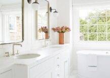 White master bathroom with white shiplap trim features curved nickel vanity mirrors flanked by black and white sconces over a honed white marble double vanity with oval sinks and a free standing bath tub.