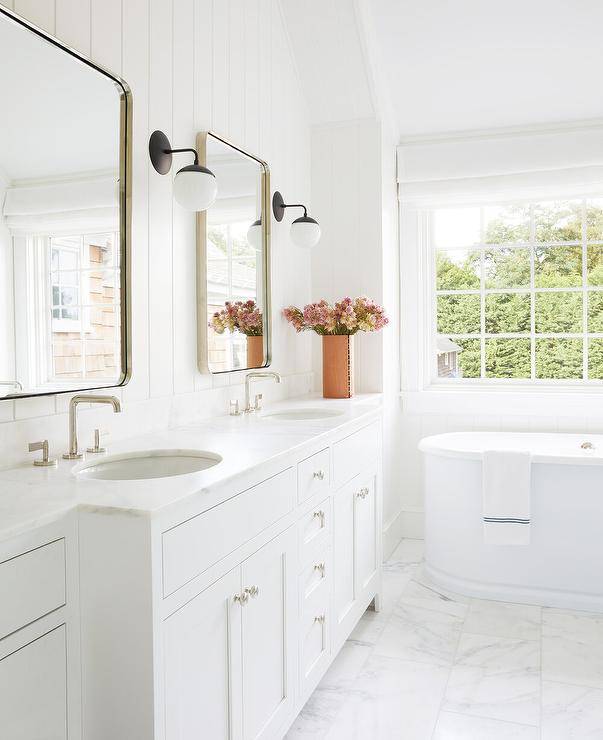 White master bathroom with white shiplap trim features curved nickel vanity mirrors flanked by black and white sconces over a honed white marble double vanity with oval sinks and a free standing bath tub.