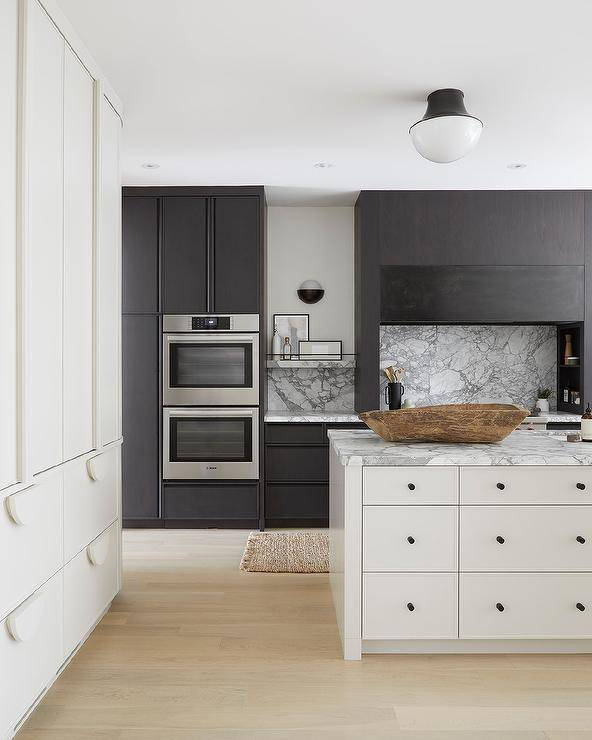 Black and white modern kitchen boasts light plank floors framing a white apothecary style island accented with a white wand gray marble countertop and matte black hardware. Behind the island, marble floating shelves are located on either side of a black cooking alcove boasting cookbook shelves flanking a white and gray marble slab backsplash.