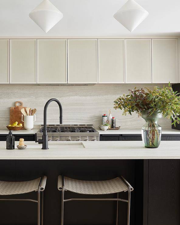 White diamond pendant lights illuminate an ivory marble countertop accenting a black kitchen island and finished with a sink with a matte black gooseneck faucet. Gorgeous backless woven gray stools sit at the island.