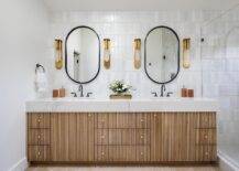 Beautifully designed bathroom boasts black oblong mirrors hung from a wall covered in light gray glazed vertical stacked tiles lit by gold sconces. The mirrors are mounted above a gold reeded double washstand adorned with brass knobs and completed with oil rubbed bronze gooseneck faucets.