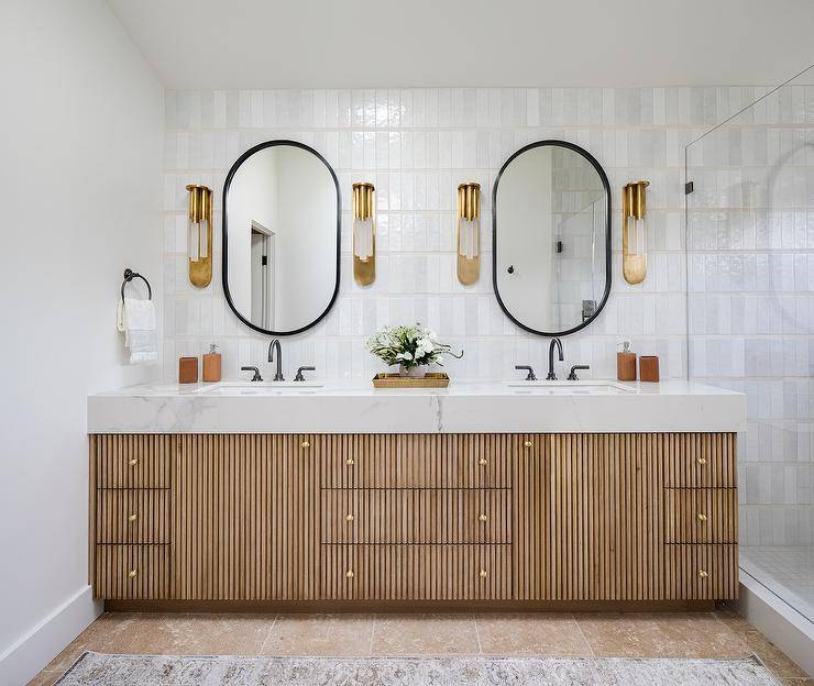 Beautifully designed bathroom boasts black oblong mirrors hung from a wall covered in light gray glazed vertical stacked tiles lit by gold sconces. The mirrors are mounted above a gold reeded double washstand adorned with brass knobs and completed with oil rubbed bronze gooseneck faucets.