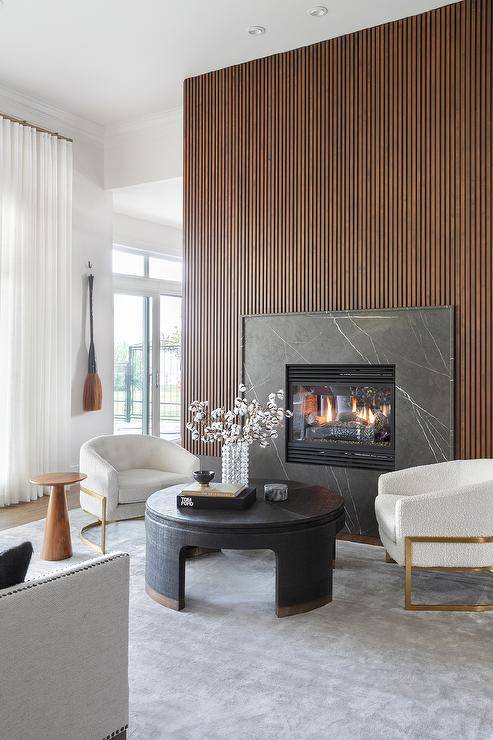 Living room features a brown reeded wall with black marble fireplace mantle, modern white and gold chairs with a round gold and black coffee table atop a gray rug and a small round accent table.