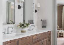 Camille Long Sconce light nickel framed vanity mirrors hung over a brown brushed oak dual washstand fitted with modern faucets and mounted against gray hexagon floor tiles. White walls are lined with gray crown moldings and gray baseboards.