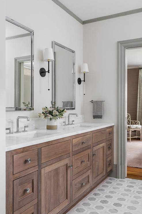 Camille Long Sconce light nickel framed vanity mirrors hung over a brown brushed oak dual washstand fitted with modern faucets and mounted against gray hexagon floor tiles. White walls are lined with gray crown moldings and gray baseboards.