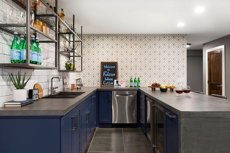 Kitchen features a blue U-shaped wet bar with concrete countertops, gray geomeric tiles backsplash, stainless steel industrial shelves on white offset tiles and large gray floor tiles.