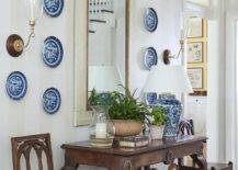 Blue decorative plates surround a foyer mirror hung from a wall clad in light gray striped wallpaper an dpositioned over a vintage console table flanked by vintage chairs. The console table is lit by a blue chinoiserie lamp and located over a wicker bin.
