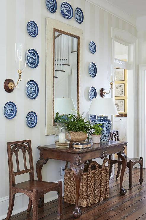 Blue decorative plates surround a foyer mirror hung from a wall clad in light gray striped wallpaper an dpositioned over a vintage console table flanked by vintage chairs. The console table is lit by a blue chinoiserie lamp and located over a wicker bin.