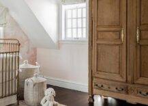 Pink French girl's nursery features a French armoire accented with antique brass hardware and placed on a dark stained wood floor beside a window covered in an off-white roman shade. A gold metal crib sits on a pink and brown rug beside white woven toy baskets.